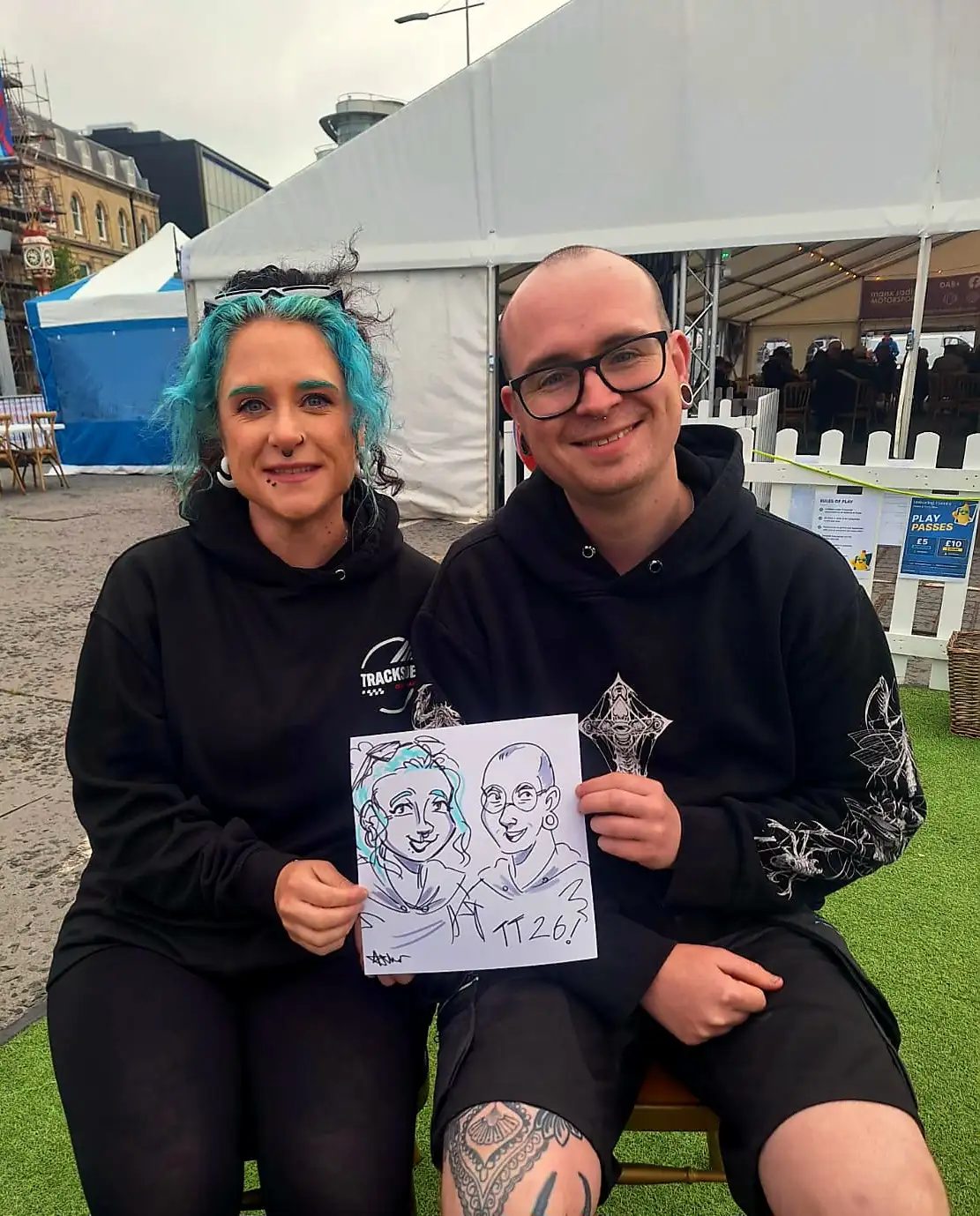 A couple holding a colourful cartoon portrait drawn during TT fortnight