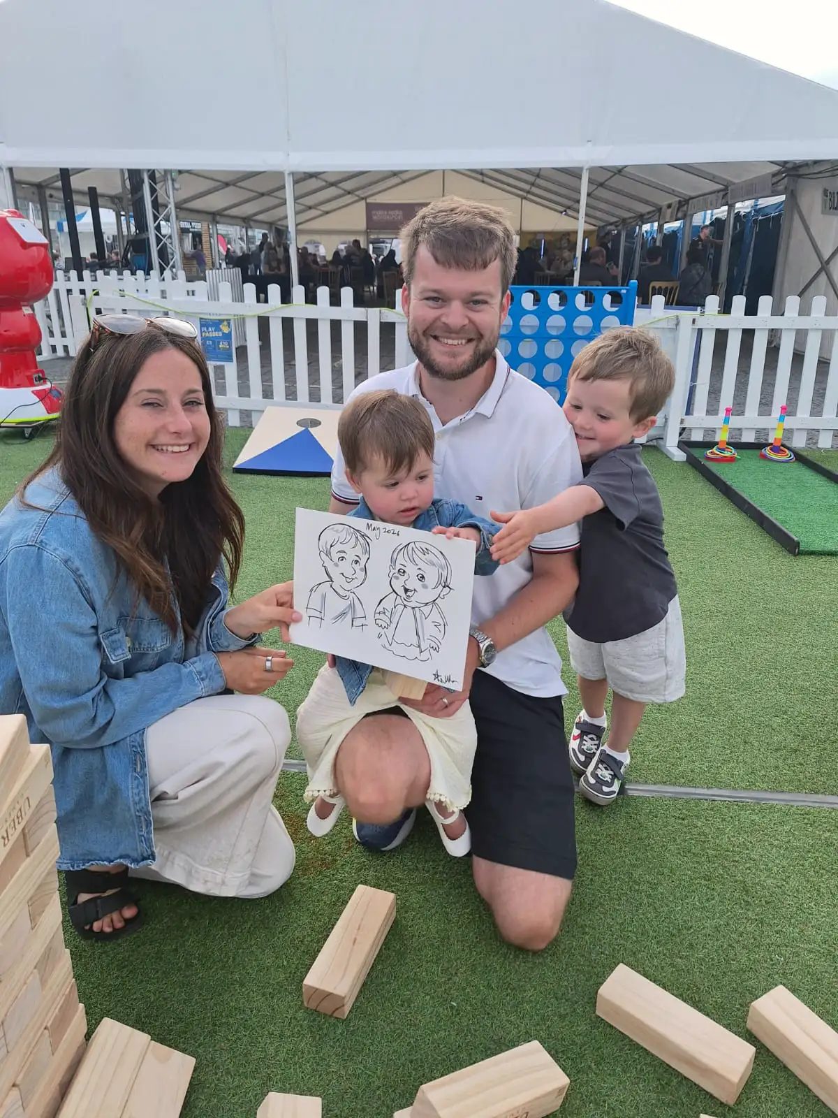 A family with two young children holding up their cartoon portrait at an outdoor event