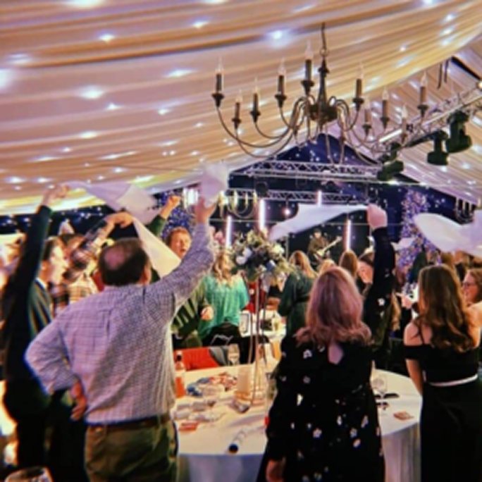 A wedding reception in full swing, guests waving napkins above their heads in a decorated marquee