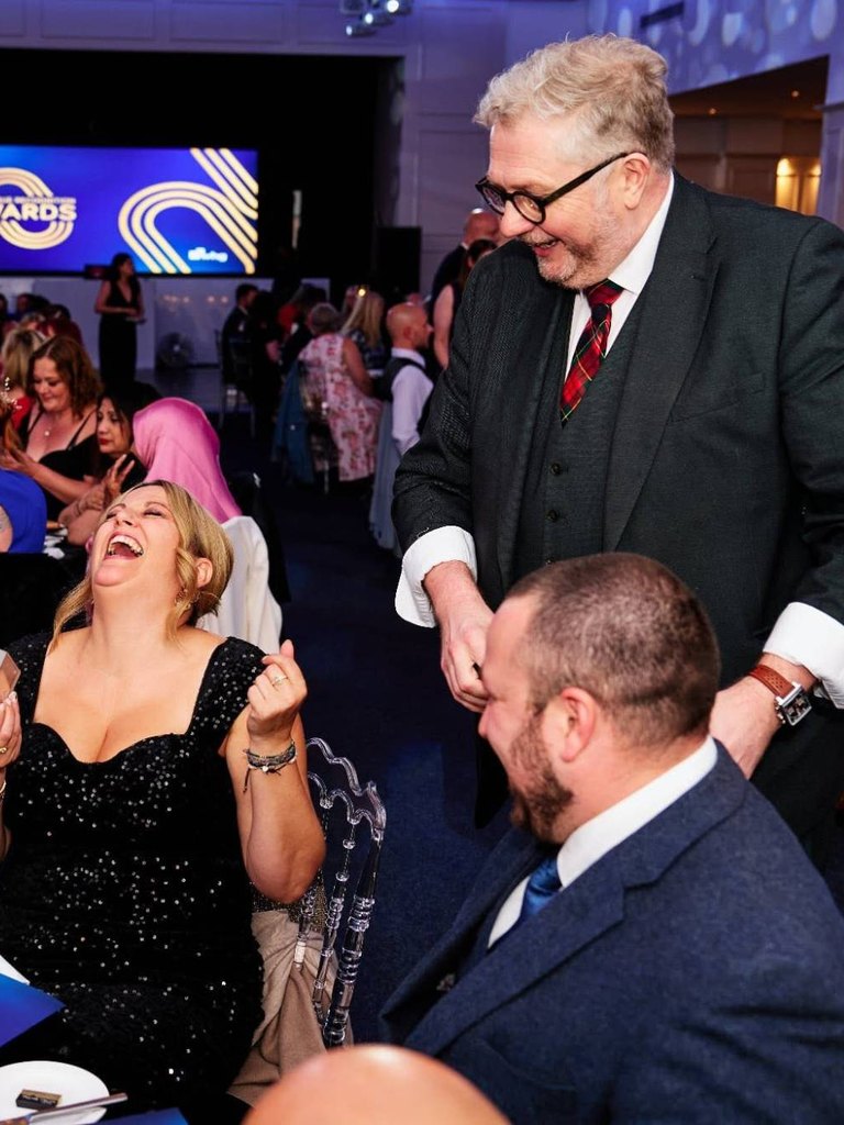 Banter Magic, a funny close-up magician from the West Midlands, in a three-piece suit and tartan tie, making a guest laugh at an awards dinner