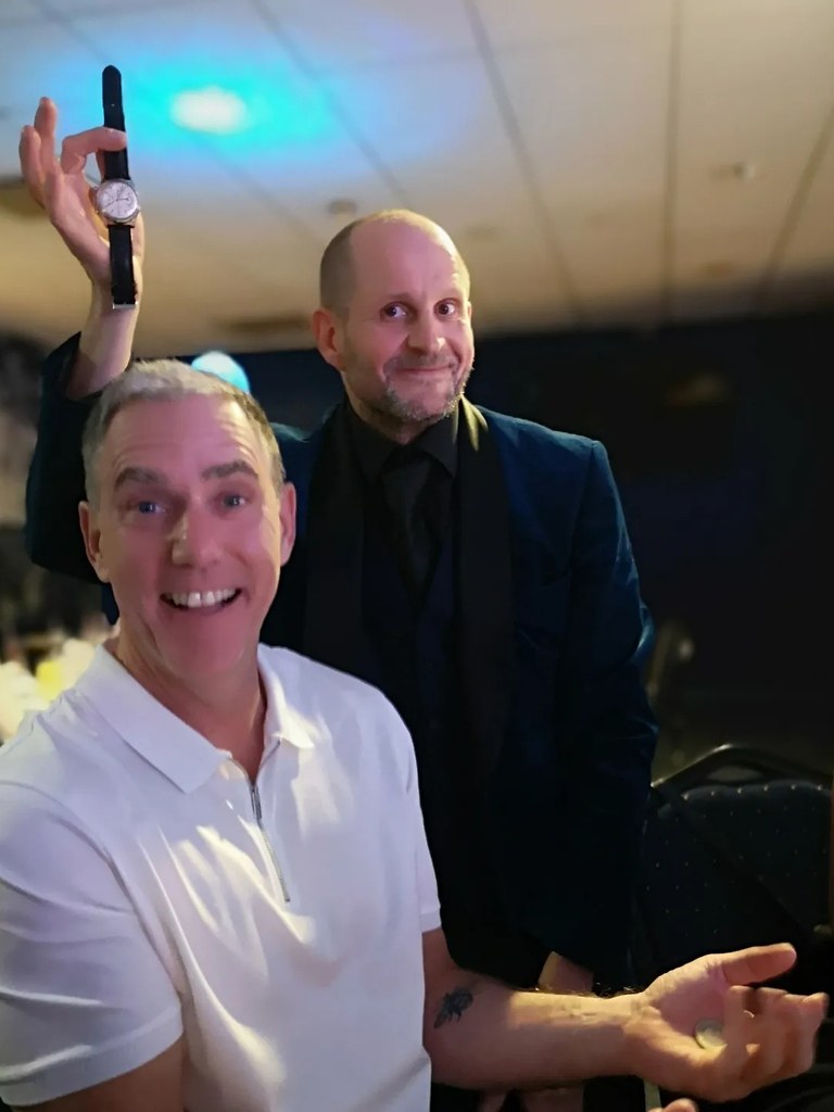 The Comedy Pickpocket holding a stolen watch in the air behind a grinning guest at an evening event
