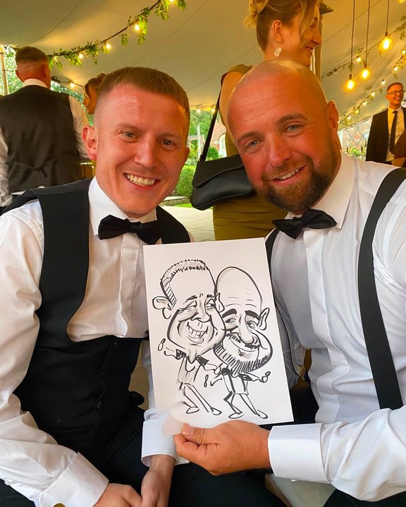 Two wedding guests in bow ties holding their caricature in a marquee