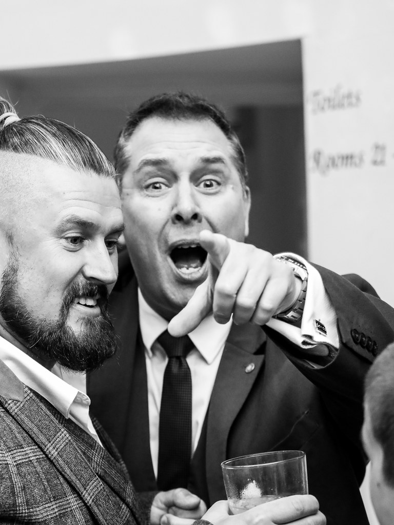 Black and white photo of the Gentleman Pickpocket pointing in mock surprise beside a guest at an evening party